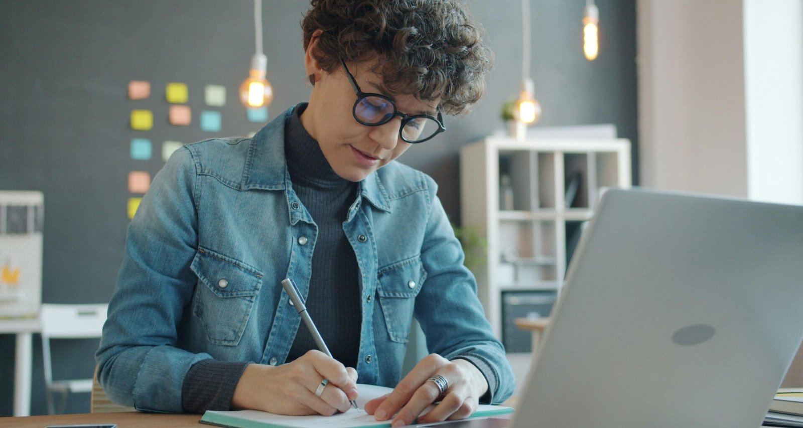 Frau mit Brille sitzt am Schreibtisch und schreibt konzentriert Notizen in ein Notizbuch, modernes Arbeitsumfeld