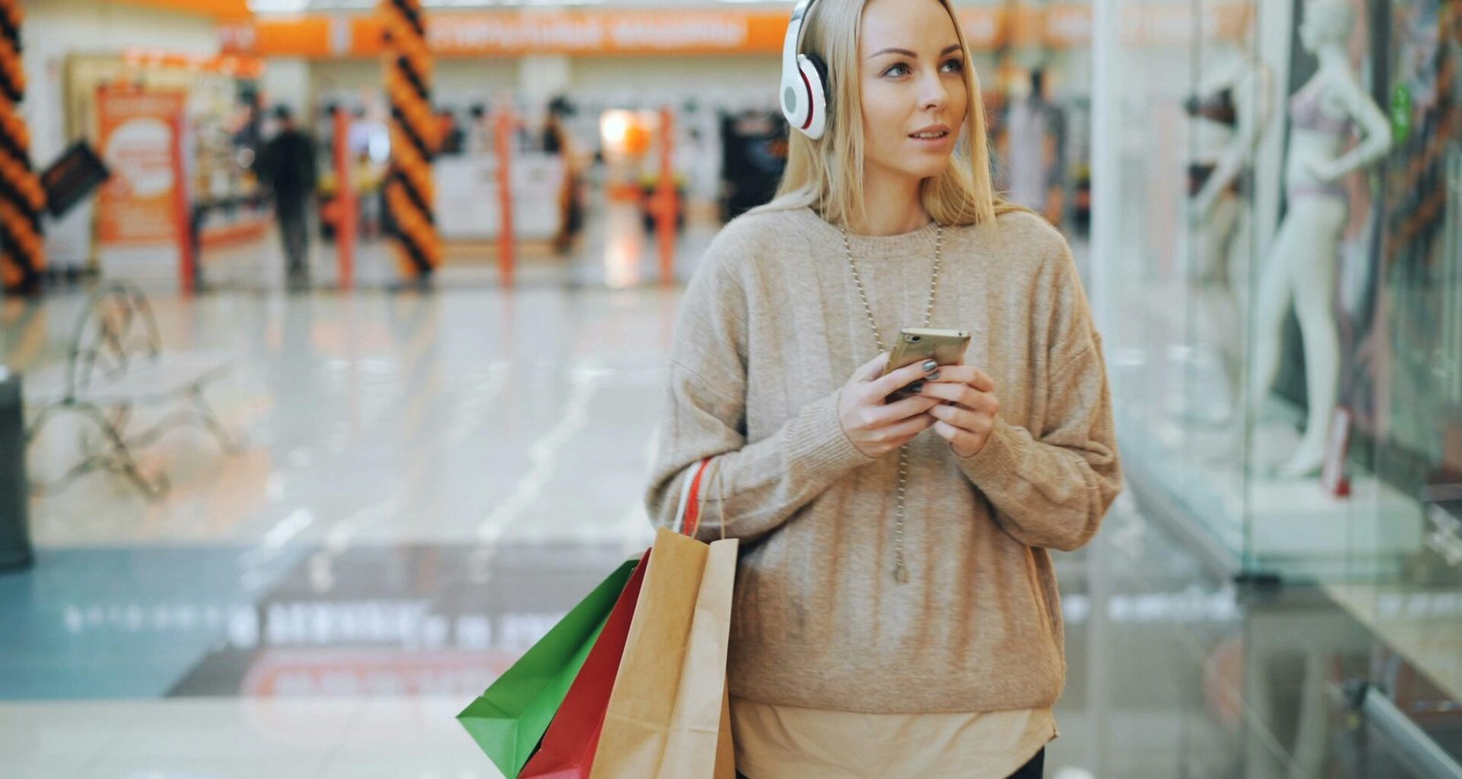 Junge Frau mit weißen Kopfhörern und Einkaufstaschen schlendert durch ein helles Einkaufszentrum