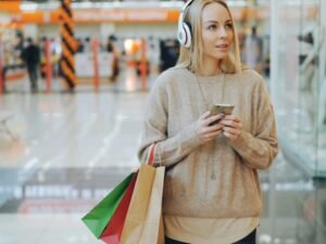 Junge Frau mit weißen Kopfhörern und Einkaufstaschen schlendert durch ein helles Einkaufszentrum