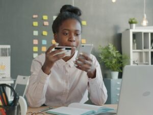 Frau am Schreibtisch hält eine Kreditkarte und ein Smartphone, Symbol für Online-Zahlungen und Digitalisierung.