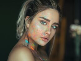 Frau mit auffälliger, mehrfarbiger Gesichtsbemalung vor neutralem Hintergrund, moderner künstlerischer Ausdruck