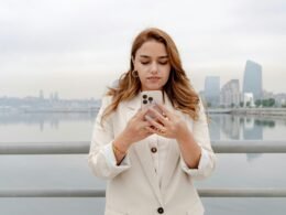 Junge Frau steht mit ihrem Smartphone vor städtischer Kulisse und nutzt ihr Handy im Freien