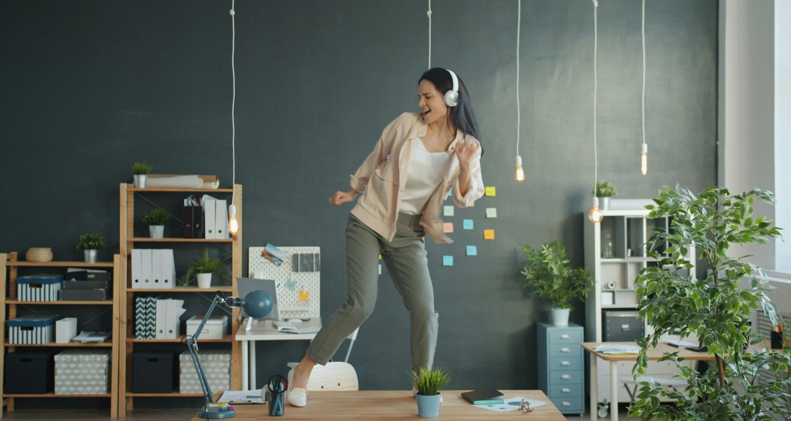 Junge Frau mit Kopfhörern tanzt auf ihrem Schreibtisch in einem modernen Büro und nutzt Musik für kreative Arbeitspausen.