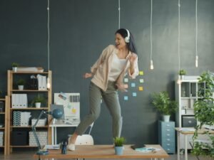 Junge Frau mit Kopfhörern tanzt auf ihrem Schreibtisch in einem modernen Büro und nutzt Musik für kreative Arbeitspausen.