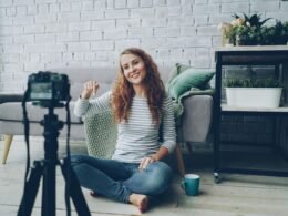 Frau winkt in die Kamera während sie ein Video in ihrem Wohnzimmer aufnimmt, Fokus auf Heimaufnahme und Content-Erstellung.