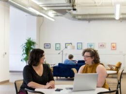 Mehrere Frauen sitzen zusammen an einem Tisch und führen ein Gespräch in einem modernen Büro oder Konferenzraum.