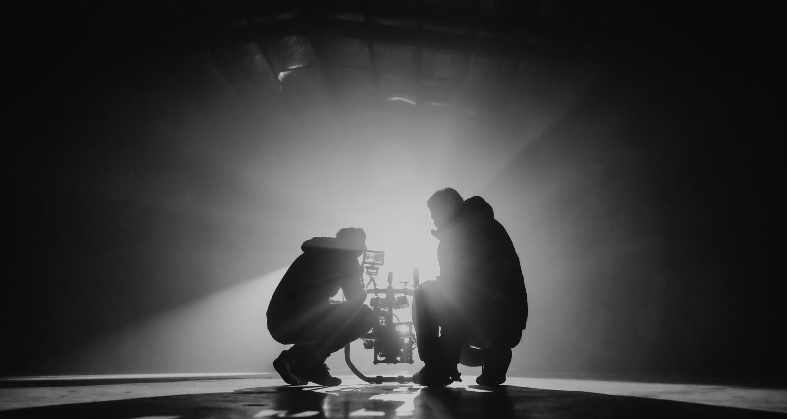 Schwarz-Weiß-Fotografie von zwei Männern, die nebeneinander kauernd auf dem Boden sitzen, aufgenommen im urbanen Umfeld