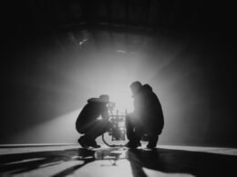 Schwarz-Weiß-Fotografie von zwei Männern, die nebeneinander kauernd auf dem Boden sitzen, aufgenommen im urbanen Umfeld