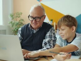 Ein älterer Mann und ein Junge sitzen nebeneinander und schauen interessiert zusammen auf einen offenen Laptop.