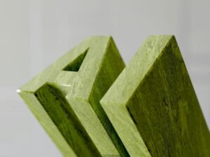 Grüne, aus Holz gefertigte Buchstaben, die das Wort „am“ auf einer hellen Oberfläche darstellen, Makroaufnahme