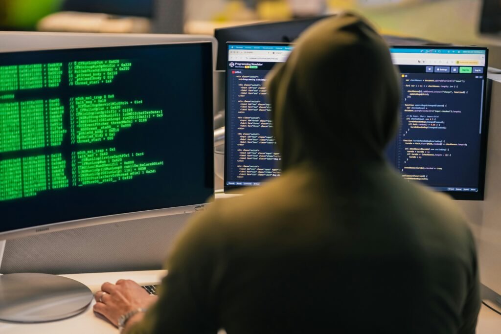 Ein Hacker mit Kapuze sitzt vor mehreren Computerbildschirmen und arbeitet an komplexen digitalen Aufgaben im Büro.