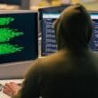 Hacker mit Hoodie arbeitet nachts an mehreren Computern und Monitoren mit Quellcode in dunkler Umgebung
