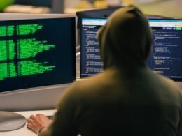 Hacker mit Kapuzenpullover analysiert Daten auf mehreren Monitoren in einem dunklen Büro