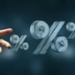 Eine Hand erreicht nach mehreren frei schwebenden Prozentzeichen vor neutralem Hintergrund – Symbolbild für Zinsen oder Rabatte.