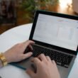 Hände tippen auf Laptop-Tastatur, auf dem Bildschirm ist eine geöffnete digitale Tabelle zu sehen