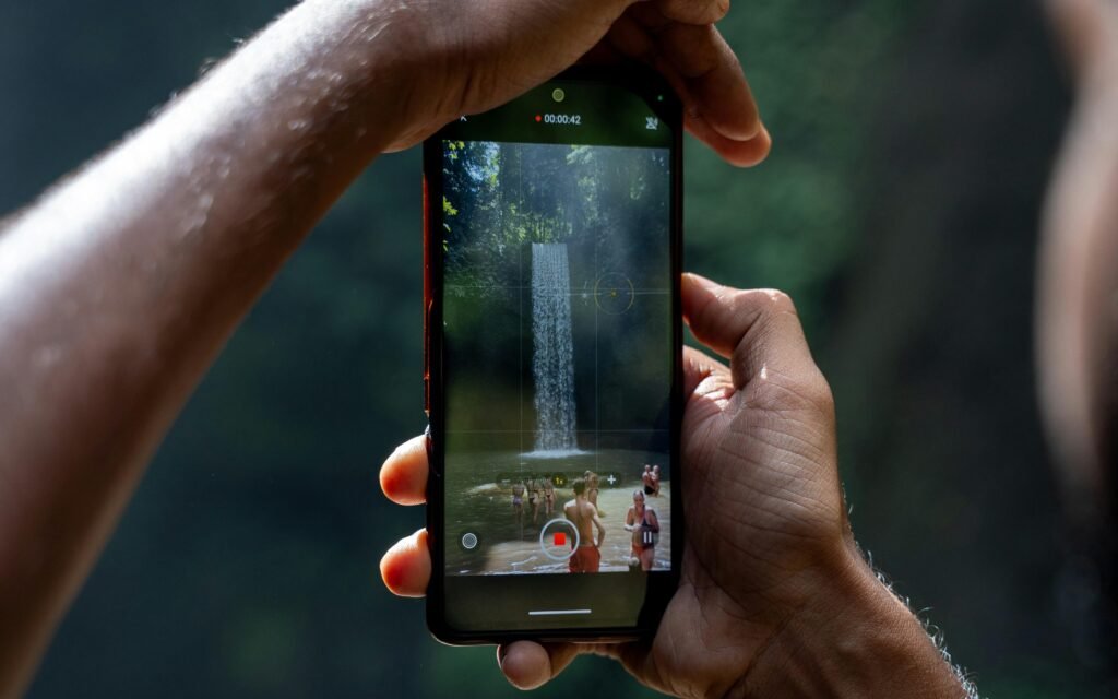 Zwei Hände halten ein Smartphone, das einen Wasserfall filmt; moderner Technologieeinsatz in der Natur