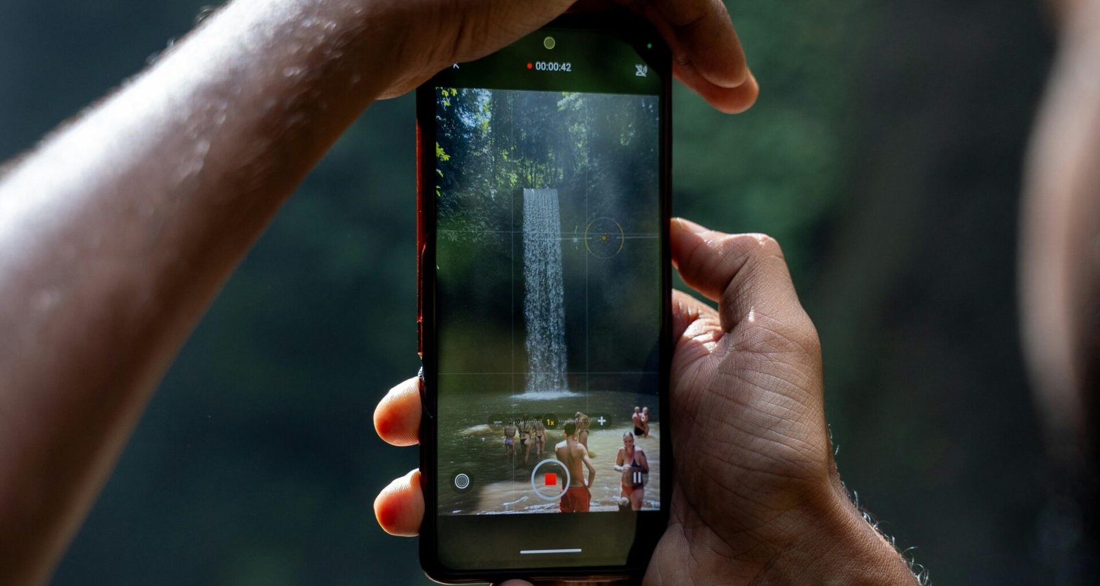 Zwei Hände halten ein Smartphone, das einen Wasserfall filmt; moderner Technologieeinsatz in der Natur