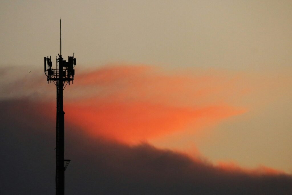 Ein Mobilfunkmast als Silhouette vor einem farbigen Sonnenuntergangshimmel, Fokus auf moderne Kommunikationsinfrastruktur.
