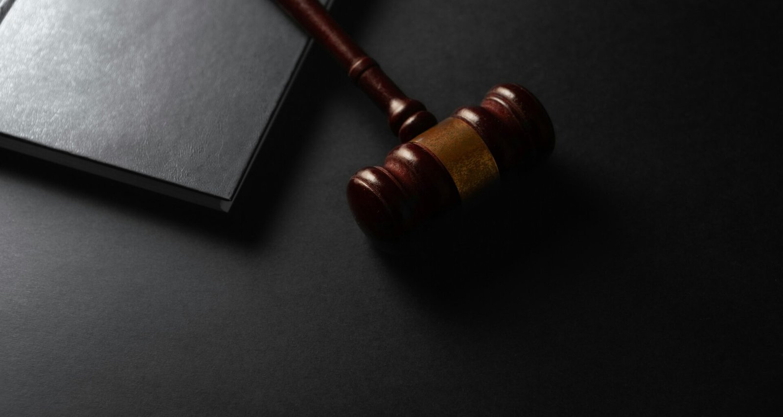 Ein hölzerner Richterhammer ruht neben einem aufgeschlagenen Buch auf dunklem Untergrund, Fokus auf Recht und Gesetz