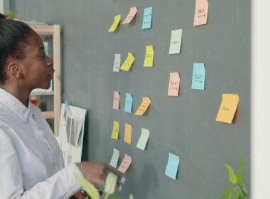 Junge Frau betrachtet eine Wand mit vielen farbigen Haftnotizen, um Aufgaben und Ideen im Team zu organisieren.