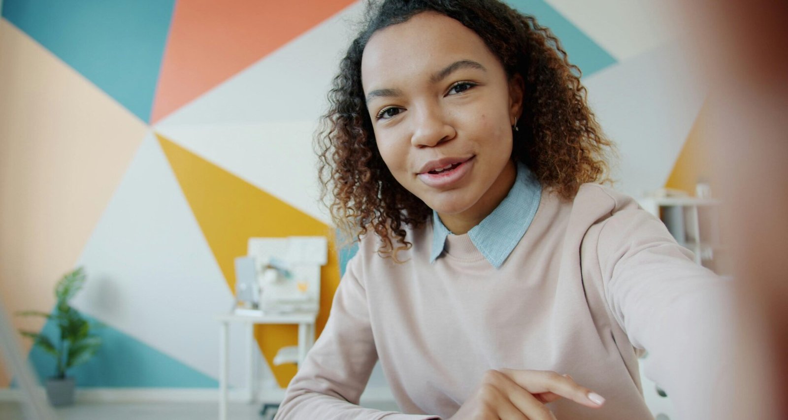 Junge Frau sitzt in einem farbenfrohen Zimmer und spricht direkt in eine Kamera, umgeben von kreativem Ambiente