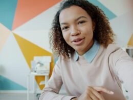 Junge Frau sitzt in einem farbenfrohen Zimmer und spricht direkt in eine Kamera, umgeben von kreativem Ambiente