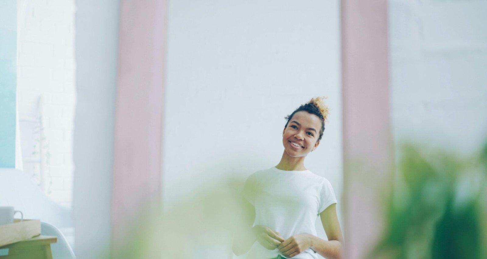 Junge Frau mit dunklen Haaren lächelt und betrachtet ihr Spiegelbild in einem hellen Raum mit neutralem Hintergrund.