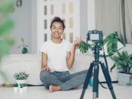 Junge Frau sitzt in einem Zimmer und nimmt sich selbst mit einer Kamera auf, vorbereitet für Videoinhalte