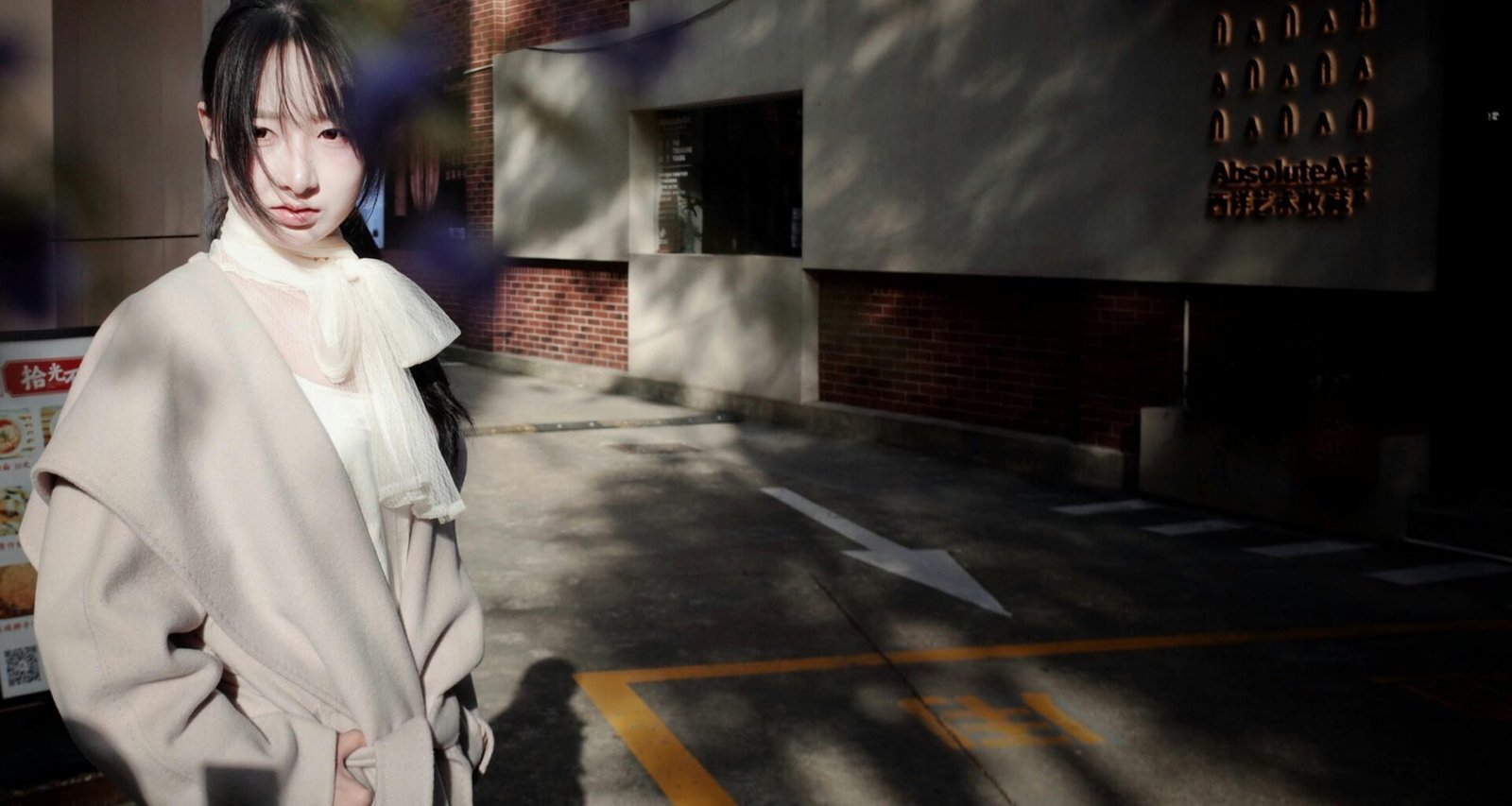 Junge Frau im hellen Mantel steht draußen neben einem Backsteingebäude, urbanes Setting, aufgenommen bei Tageslicht