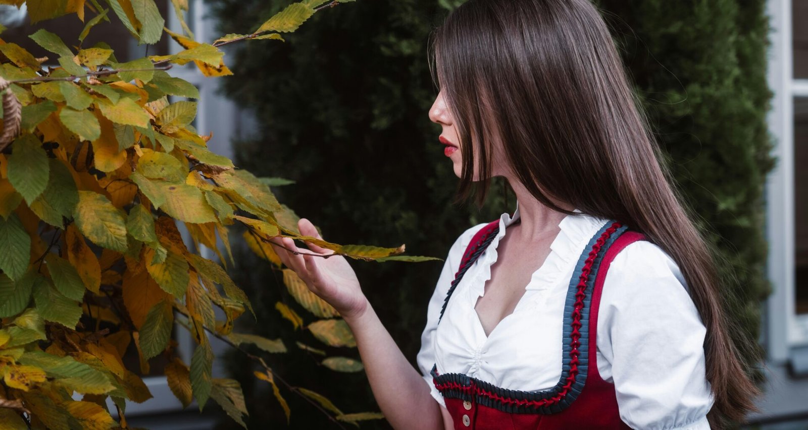 Junge Frau in traditioneller Kleidung berührt herbstliche Blätter an einem Baum vor natürlichem Hintergrund