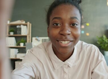 Junge Frau sitzt lächelnd an einem Schreibtisch im hellen Büro mit Laptop und Notizbuch vor sich