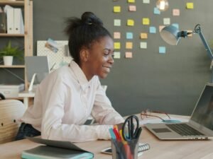 Junge Frau sitzt lächelnd am Schreibtisch und arbeitet am Laptop, modernes Büro, Tageslicht