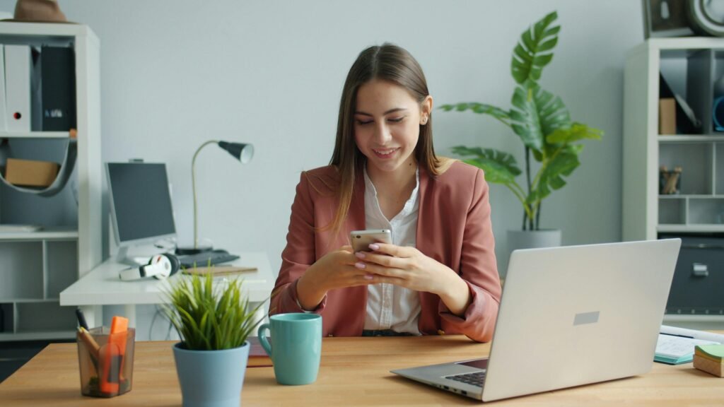 Junge Frau sitzt am Schreibtisch und lächelt, während sie mit ihrem Smartphone arbeitet; modernes Arbeitsumfeld.