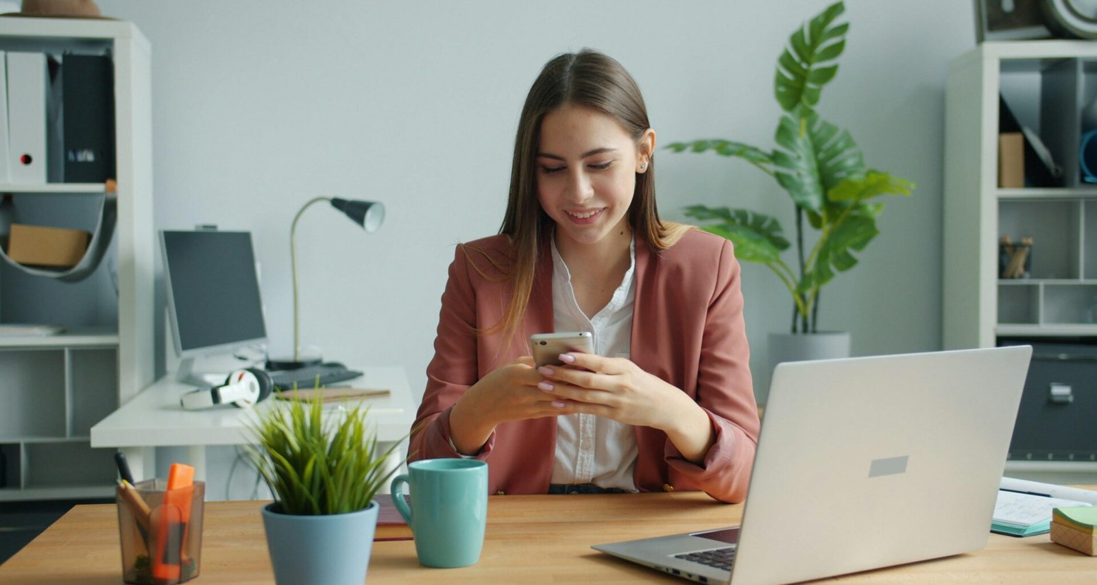 Junge Frau sitzt am Schreibtisch und lächelt, während sie mit ihrem Smartphone arbeitet; modernes Arbeitsumfeld.