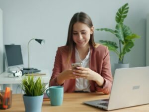 Junge Frau sitzt am Schreibtisch und lächelt, während sie mit ihrem Smartphone arbeitet; modernes Arbeitsumfeld.