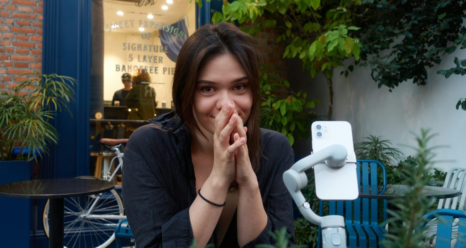 Junge Frau sitzt und lächelt, ihre Hände sind vor dem Gesicht gefaltet, neutraler Hintergrund