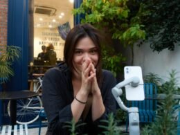 Junge Frau sitzt und lächelt, ihre Hände sind vor dem Gesicht gefaltet, neutraler Hintergrund