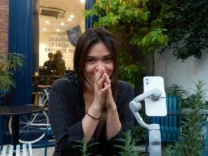 Junge Frau sitzt und lächelt, ihre Hände sind vor dem Gesicht gefaltet, neutraler Hintergrund