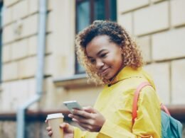 Junge Frau mit Kaffee in der Hand und Smartphone beim Gehen im Freien, im urbanen Umfeld fotografiert