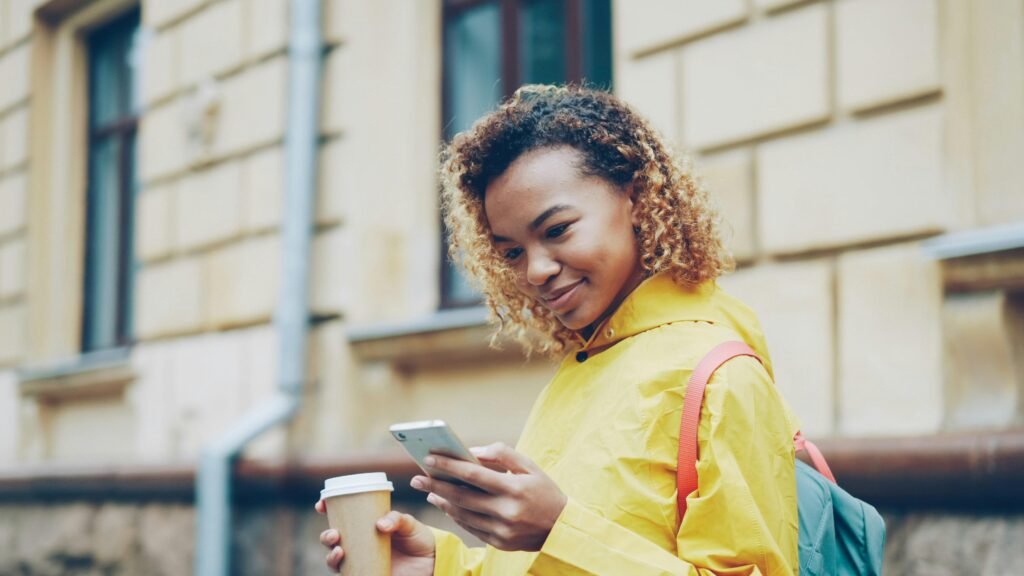 Junge Frau geht draußen mit Kaffeebecher in der Hand und blickt auf ihr Smartphone, alltägliche urbane Szene