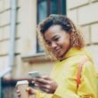 Junge Frau geht draußen mit Kaffeebecher in der Hand und blickt auf ihr Smartphone, alltägliche urbane Szene