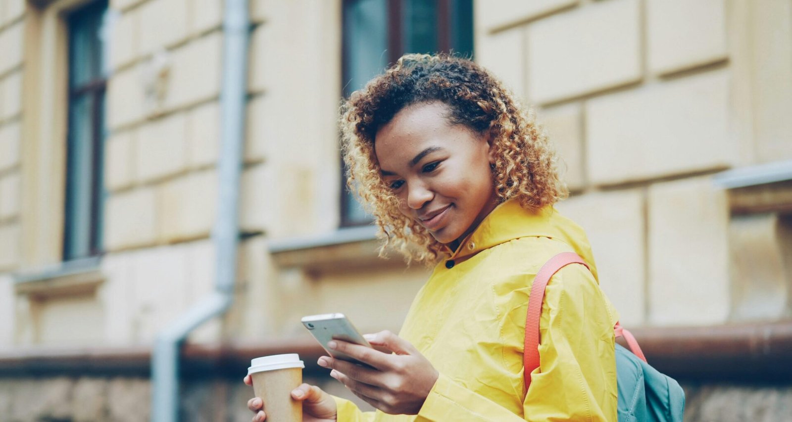 Junge Frau geht draußen mit Kaffeebecher in der Hand und blickt auf ihr Smartphone, alltägliche urbane Szene