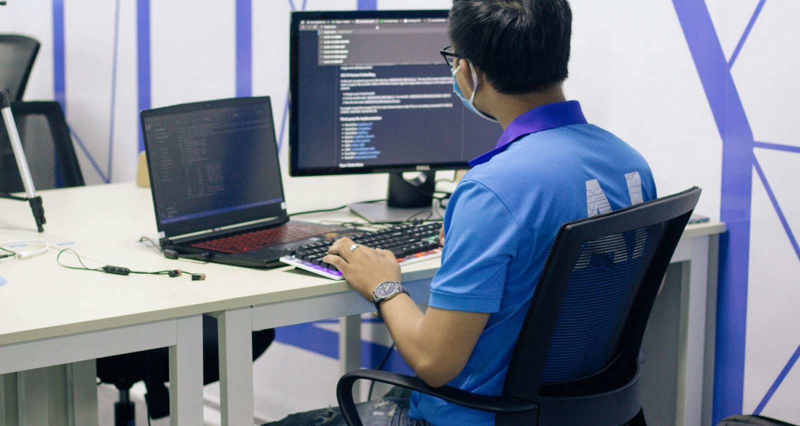 Junge Person im blauen T-Shirt sitzt auf einem schwarzen Drehstuhl und arbeitet am Computer in einem modernen Büro