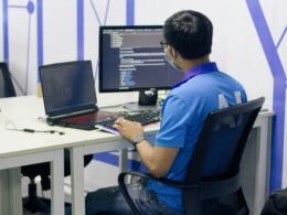 Junge Person im blauen T-Shirt sitzt auf einem schwarzen Drehstuhl und arbeitet am Computer in einem modernen Büro