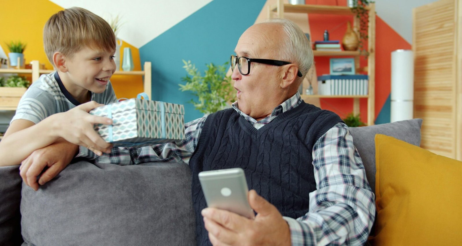 Ein Junge gibt einem älteren Mann, vermutlich seinem Großvater, ein in Geschenkpapier verpacktes Präsent zu Hause.