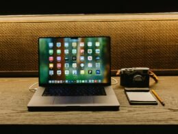 Laptop, Kamera und Notizblock liegen auf einem hölzernen Schreibtisch, bereit für kreative und digitale Arbeitsprozesse.