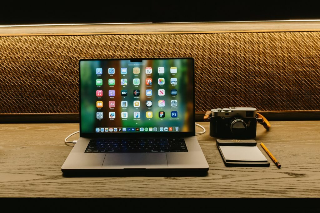 Laptop, Kamera und Notizbuch arrangiert auf einem hölzernen Schreibtisch als Arbeitsumgebung für digitale Projekte.