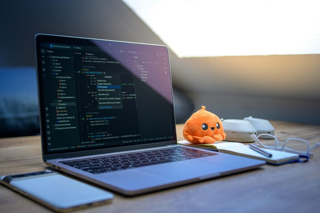 Ein Schreibtisch mit aufgeklapptem Laptop, sichtbarem Code, Smartphone, Brille und einem Plüschtier im Arbeitsumfeld