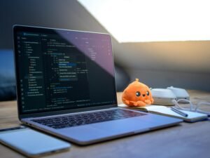 Laptop mit geöffnetem Code-Editor, daneben Smartphone, Brille und ein Plüschtier – ideal für Artikel zu Webentwicklung oder Programmierung.