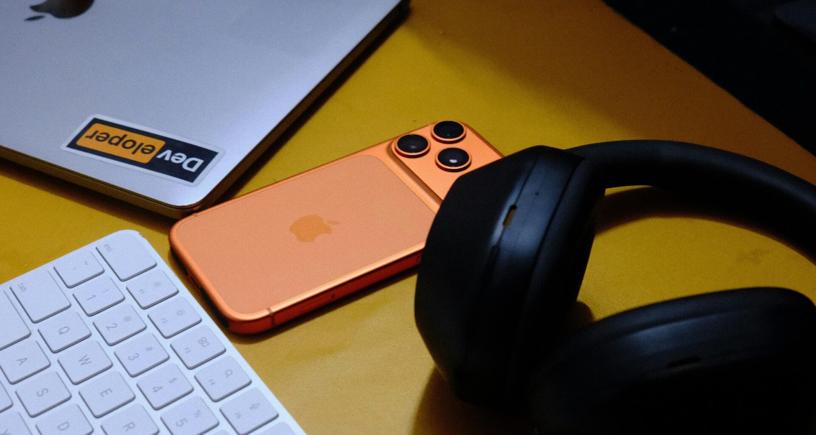 Laptop, Smartphone, Tastatur und Kopfhörer nebeneinander auf einer gelben Fläche arrangiert, Fokus auf technischen Geräten